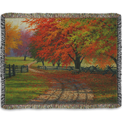A woven throw blanket designed with a painting of a road winding through a fenced field, surrounded by multicolored fall maple trees.