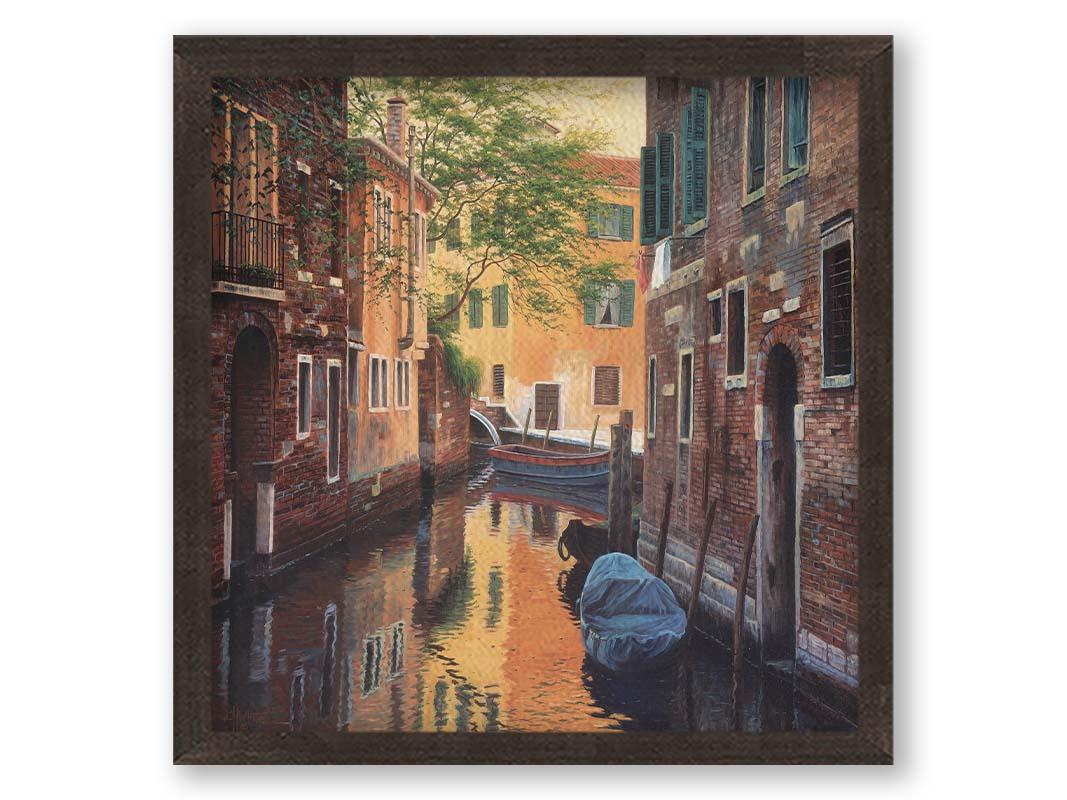 A painting of a canal alleyway between old brick buildings in Venice, Italy, during the day. The buildings reflect off the water in which a few small wood boats float. Printed on canvas and framed.