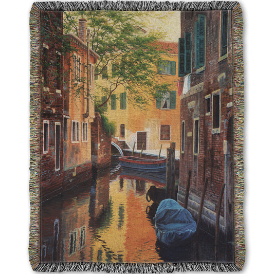 A woven throw blanket designed with a painting of a canal alleyway between old brick buildings in Venice, Italy, during the day. The buildings reflect off the water in which a few small wood boats float.