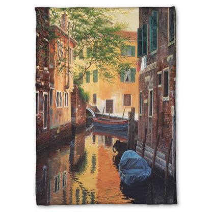 A fleece throw blanket designed with a painting of a canal alleyway between old brick buildings in Venice, Italy, during the day. The buildings reflect off the water in which a few small wood boats float.