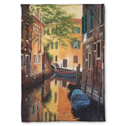 A fleece throw blanket designed with a painting of a canal alleyway between old brick buildings in Venice, Italy, during the day. The buildings reflect off the water in which a few small wood boats float.