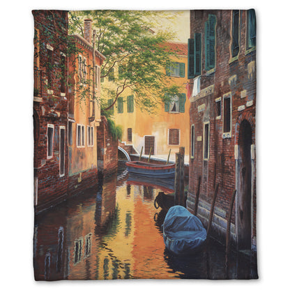 A fleece throw blanket designed with a painting of a canal alleyway between old brick buildings in Venice, Italy, during the day. The buildings reflect off the water in which a few small wood boats float.