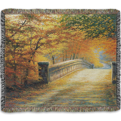 A woven throw blanket designed with a painting looking down a bridge road during fall. The trees lining the road have turned yellow and orange for autumn, and some of their fallen leaves are scatted along the path.