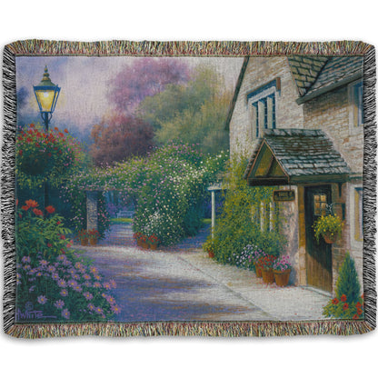 A woven throw blanket designed with a painting of a bed and breakfast storefront, with a streetlamp-lined path leading to the garden full of blooming flowers.