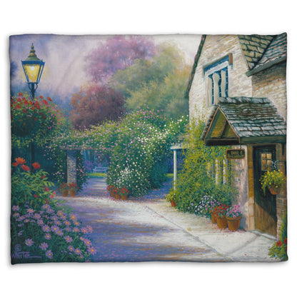 A fleece throw blanket designed with a painting of a bed and breakfast storefront, with a streetlamp-lined path leading to the garden full of blooming flowers.