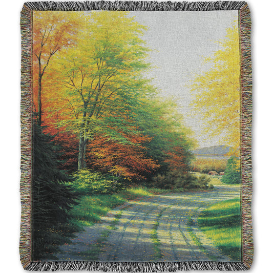 A woven throw blanket designed with a painting looking down a country road, lined with both deciduous and coniferous trees. Several of the trees' leaves have turned orange and yellow due to fall.