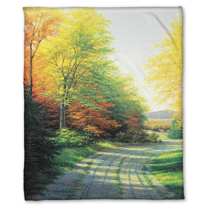 A fleece throw blanket designed with a painting looking down a country road, lined with both deciduous and coniferous trees. Several of the trees' leaves have turned orange and yellow due to fall.