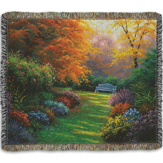 A woven throw blanket designed with a painting of a garden path during fall. The trees surrounding the garden have turned orange and yellow. A grassy path through the garden is surrounded by bushes of flowers in every color, and a bench can be seen up ahead.