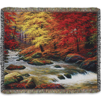 A woven throw blanket designed with a painting of a river through an autumn forest, with red and yellow trees and an orange, leaf-coated ground.