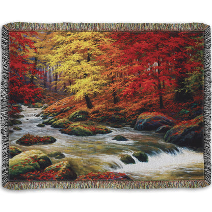 A woven throw blanket designed with a painting of a river through an autumn forest, with red and yellow trees and an orange, leaf-coated ground.