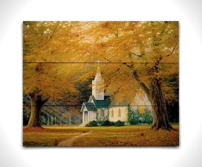 A painting of a white church in a forest of yellow and orange trees. Deer roam in the leaf-coated yard, and sun beams shine through the trees to highlight the building. Printed on a wood pallet.