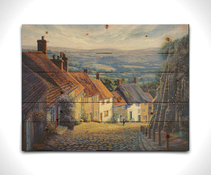 A painting looking down a cobblestone road on a hill lined with houses. A person can be seen walking down the path past flower-laden walls. Printed on a wood pallet.