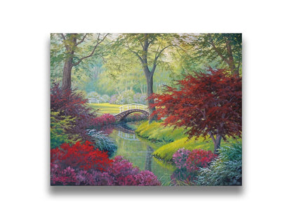 A painting of a garden park, with a stone bridge crossing a stream surrounded by flowering plants. Red, pink, and white flowers stand out against the vibrant greens of the grasses and trees. A white bench can be seen in a grassy clearing in the distance. Printed on canvas.