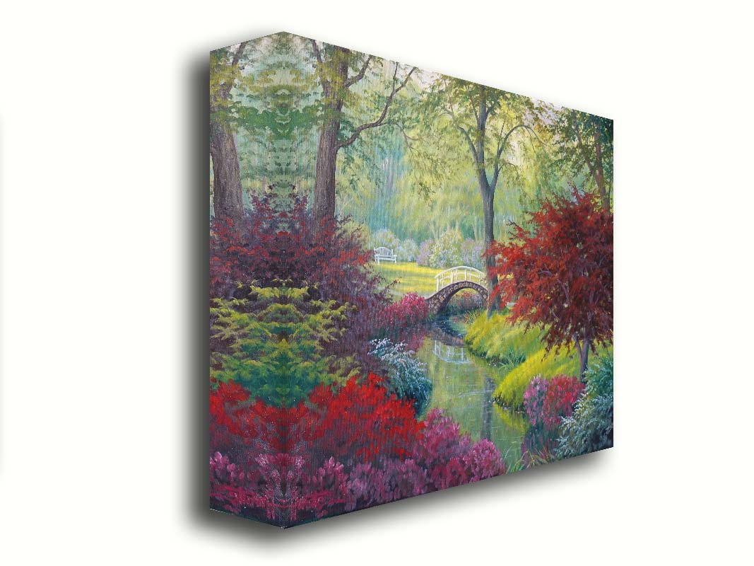 A painting of a garden park, with a stone bridge crossing a stream surrounded by flowering plants. Red, pink, and white flowers stand out against the vibrant greens of the grasses and trees. A white bench can be seen in a grassy clearing in the distance. Printed on canvas.