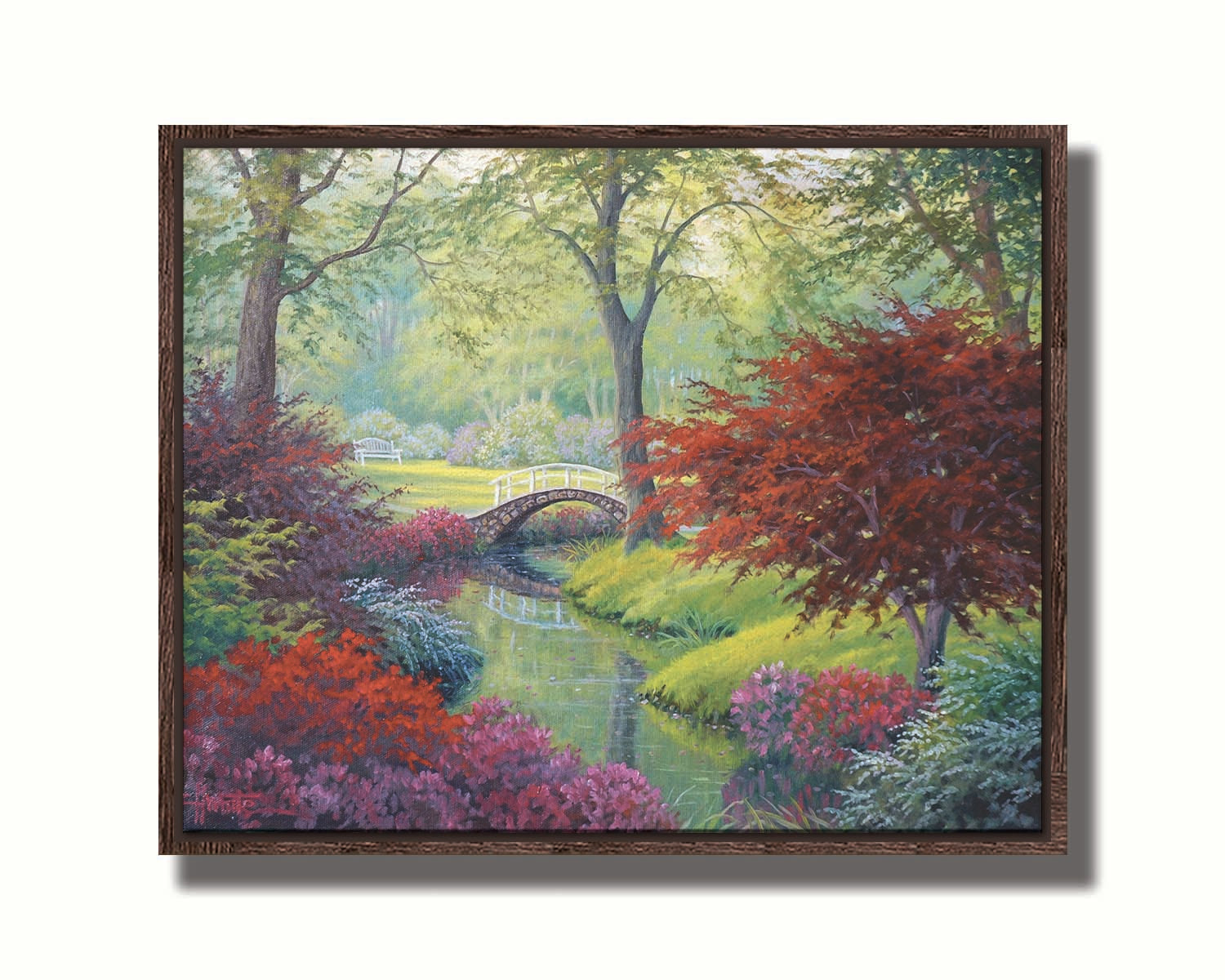 A painting of a garden park, with a stone bridge crossing a stream surrounded by flowering plants. Red, pink, and white flowers stand out against the vibrant greens of the grasses and trees. A white bench can be seen in a grassy clearing in the distance. Printed on canvas in a float frame.