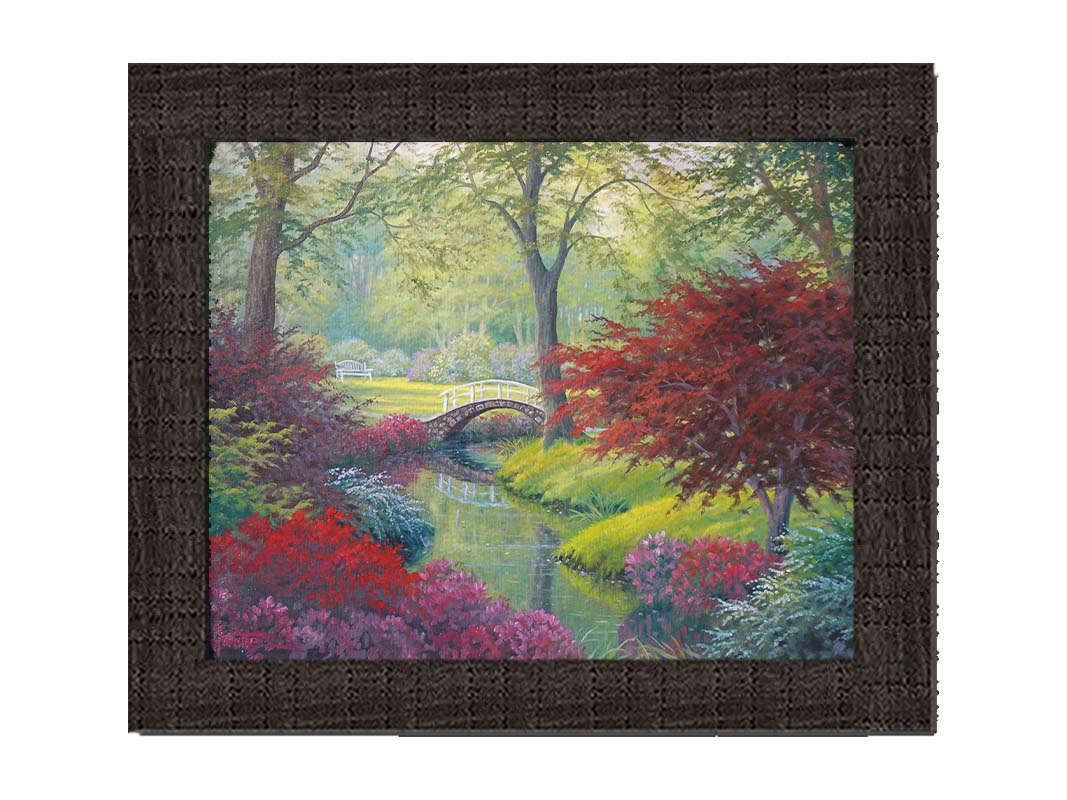 A painting of a garden park, with a stone bridge crossing a stream surrounded by flowering plants. Red, pink, and white flowers stand out against the vibrant greens of the grasses and trees. A white bench can be seen in a grassy clearing in the distance. Printed on canvas and framed.