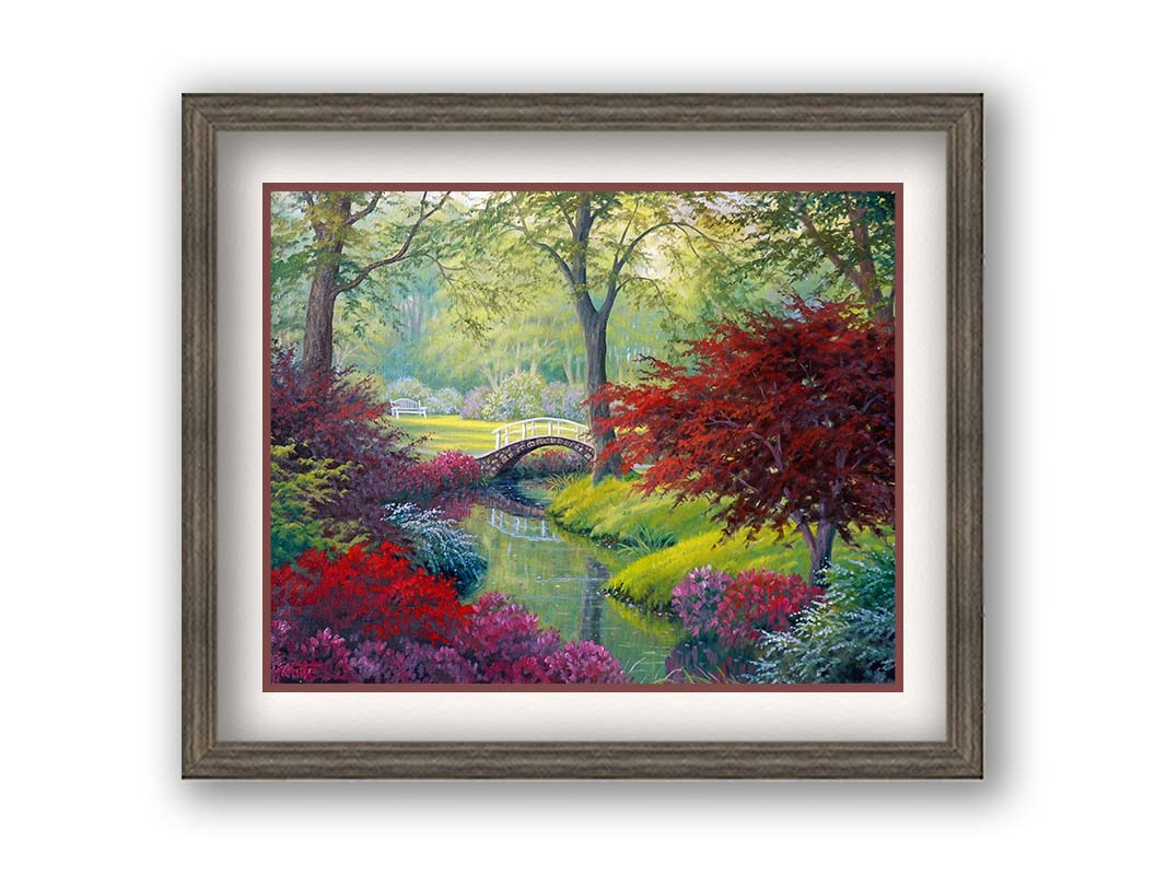 A painting of a garden park, with a stone bridge crossing a stream surrounded by flowering plants. Red, pink, and white flowers stand out against the vibrant greens of the grasses and trees. A white bench can be seen in a grassy clearing in the distance. Printed on paper, matted, and framed.