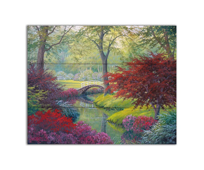 A painting of a garden park, with a stone bridge crossing a stream surrounded by flowering plants. Red, pink, and white flowers stand out against the vibrant greens of the grasses and trees. A white bench can be seen in a grassy clearing in the distance. Printed on a box board.