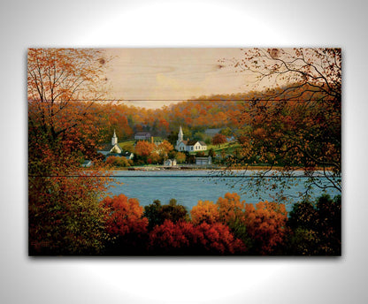 A painting of a small lakeside town, surrounded by an autumn forest. Orange and red leaves contrast green grass and vibrant blue water. Printed on a wood pallet.