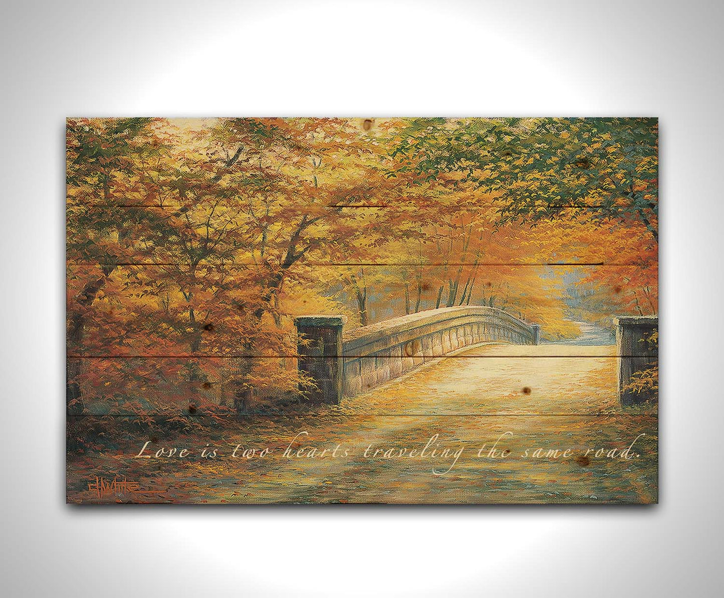 A painting looking down a bridge road during fall. The trees lining the road have turned yellow and orange for autumn, and some of their fallen leaves are scatted along the path. The quote "Love is two hearts traveling the same road" is overlayed across the image in white. Printed on a wood pallet.