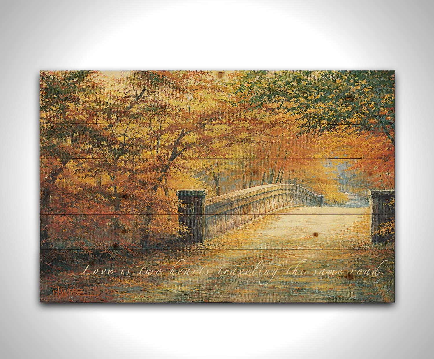 A painting looking down a bridge road during fall. The trees lining the road have turned yellow and orange for autumn, and some of their fallen leaves are scatted along the path. The quote "Love is two hearts traveling the same road" is overlayed across the image in white. Printed on a wood pallet.