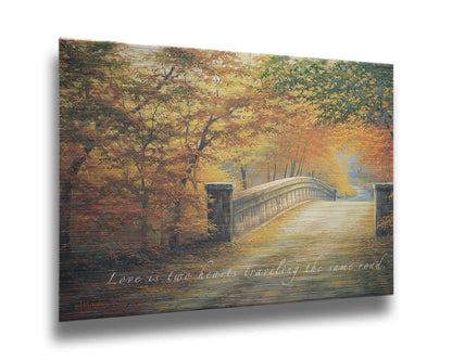 A painting looking down a bridge road during fall. The trees lining the road have turned yellow and orange for autumn, and some of their fallen leaves are scatted along the path. The quote "Love is two hearts traveling the same road" is overlayed across the image in white. Printed on metal.