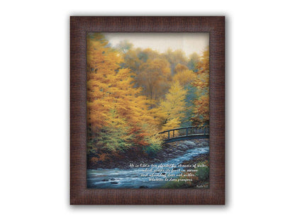 A painting of a forest stream, passing under an arched bridge. The trees are changing to yellow and orange for the fall season. The bottom right corner is overlaid with Psalm 1:3 in the Bible, reading "Psalm 1:3 from the Bible, reading "He is like a tree planted by streams of water, which yields its fruit in season and whose leaf does not wither. Whatever he does prospers." Printed on canvas and framed.