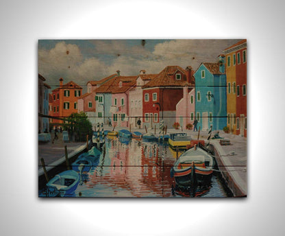 A painting of a canal in the Murano islands in Italy. Several small boats are docked along the sides, and bright, colorful buildings line the paths. Printed on a wood pallet.