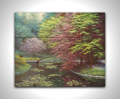 A painting of a peaceful garden pond covered in lily pads, and surrounded by vibrant red and yellow flowers, striking red maples, and blossoming white and pink trees. Printed on a wood pallet.