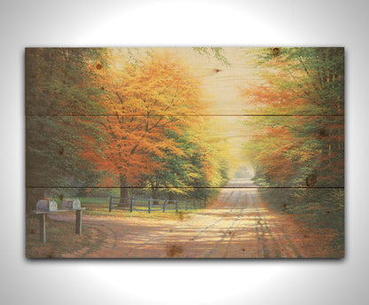 A painting of a gravel road, leading past a driveway with mailboxes and a quaint fence. All sides of the road are lined with trees, many of whose leaves have turned yellow and orange for fall. Printed on a wood pallet.