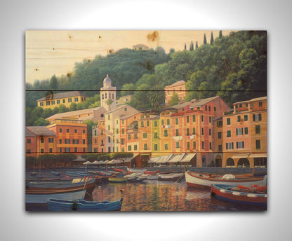 A painting of the colorful fishing town of Portofino. Many small boats are docked in the waters in the foreground, and the steep hills in the background are lush with trees. Printed on a wood pallet.