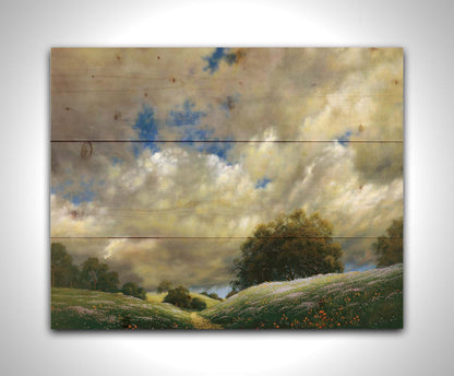 A painting of the sky over gentle hills covered in flowery meadows. Huge clouds fill the sky over the scene. Printed on a wood pallet.