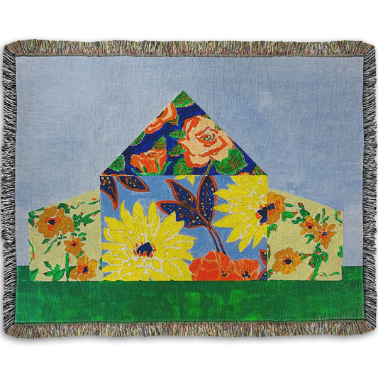 A woven throw blanket designed with an abstract painting of a barn silhouette formed of flower patterns, sitting on a green ground plane against a pale blue sky.