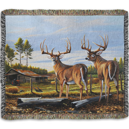 A woven throw blanket designed with a painting of a farm landscape, featuring two deer in the foreground. Fields, buildings, hay bales, and farming equipment can be seen beyond the fence, and fallen leaves cover the ground. 