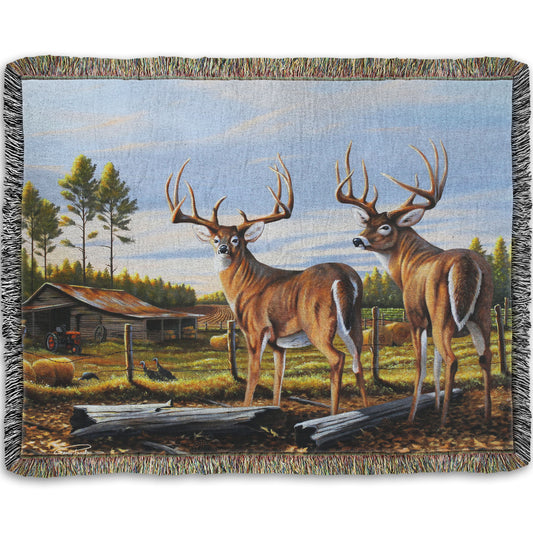 A woven throw blanket designed with a painting of a farm landscape, featuring two deer in the foreground. Fields, buildings, hay bales, and farming equipment can be seen beyond the fence, and fallen leaves cover the ground. 