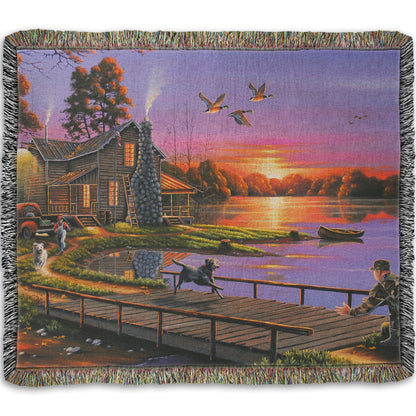 A woven throw blanket designed with a painting of a soldier returning home to his house on the lake, being greeted by his family and dogs. Geese fly overhead into the sunset. 