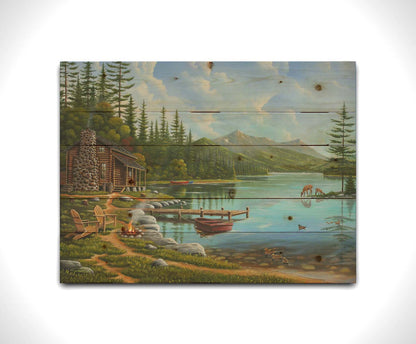 A painting of a lakeside cabin on a sunny day. Boats can be seen on the water, which reflects the mountains in the distance. Deer drink from the lake, where ducks are swimming. Wood chairs surround a small campfire on the shore nearby. Printed on a wood pallet.