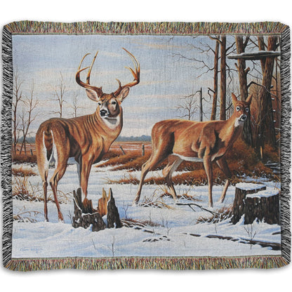 Woven throw blanket designed with deer walking through the snow between the woods and a field.