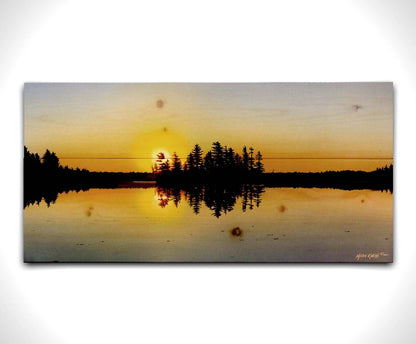 A painting of a low sun reflecting off the waters of a lake, surrounded by forest. Printed on a wood pallet.