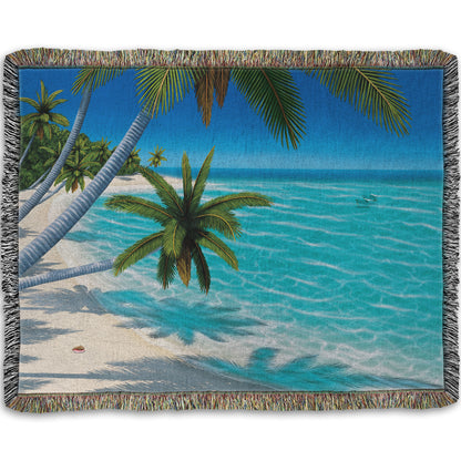 A woven throw blanket designed with a painting of a white sand beach with growing palm trees and conch shells. Sea turtles swim through the clear blue waters.