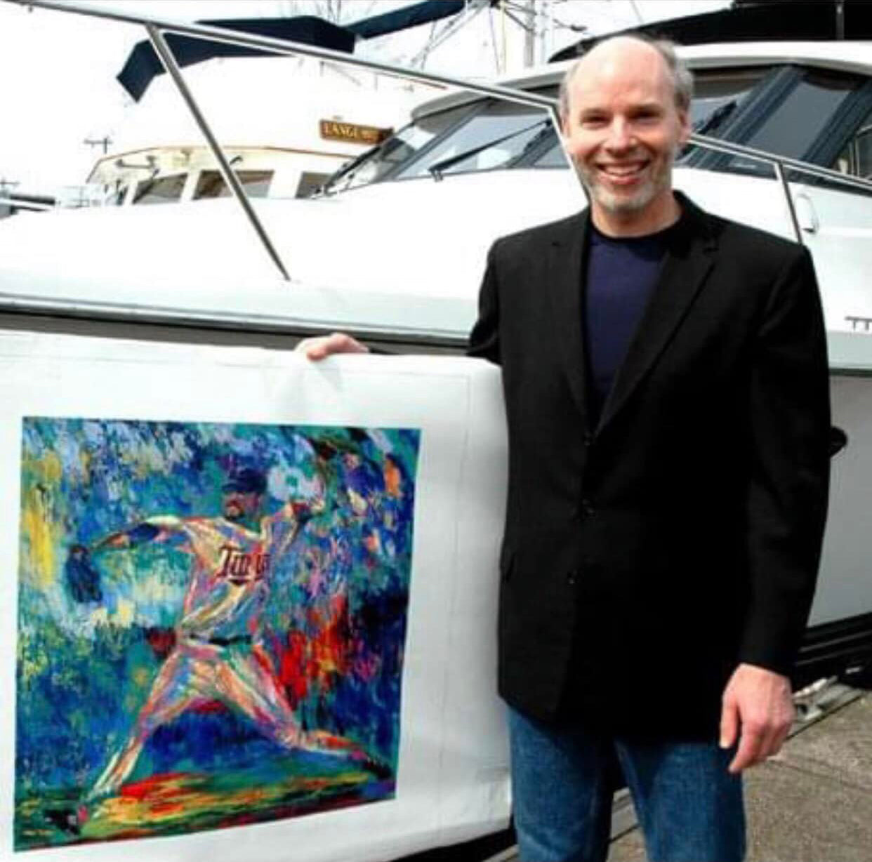 Artist Robert Blehert standing on a boat dock, holding a painting he created of baseball player Carlos Santana.