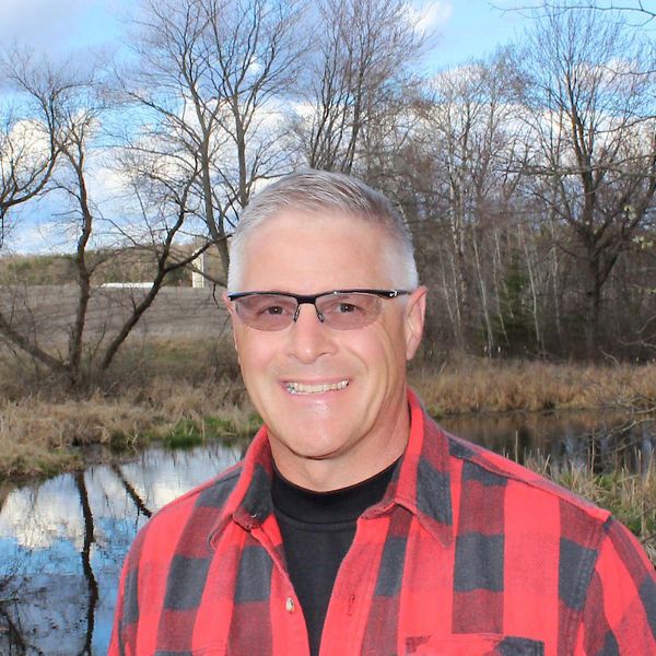 A headshot of artist Robert Andrea in front of a pond outdoors, smiling a the camera.