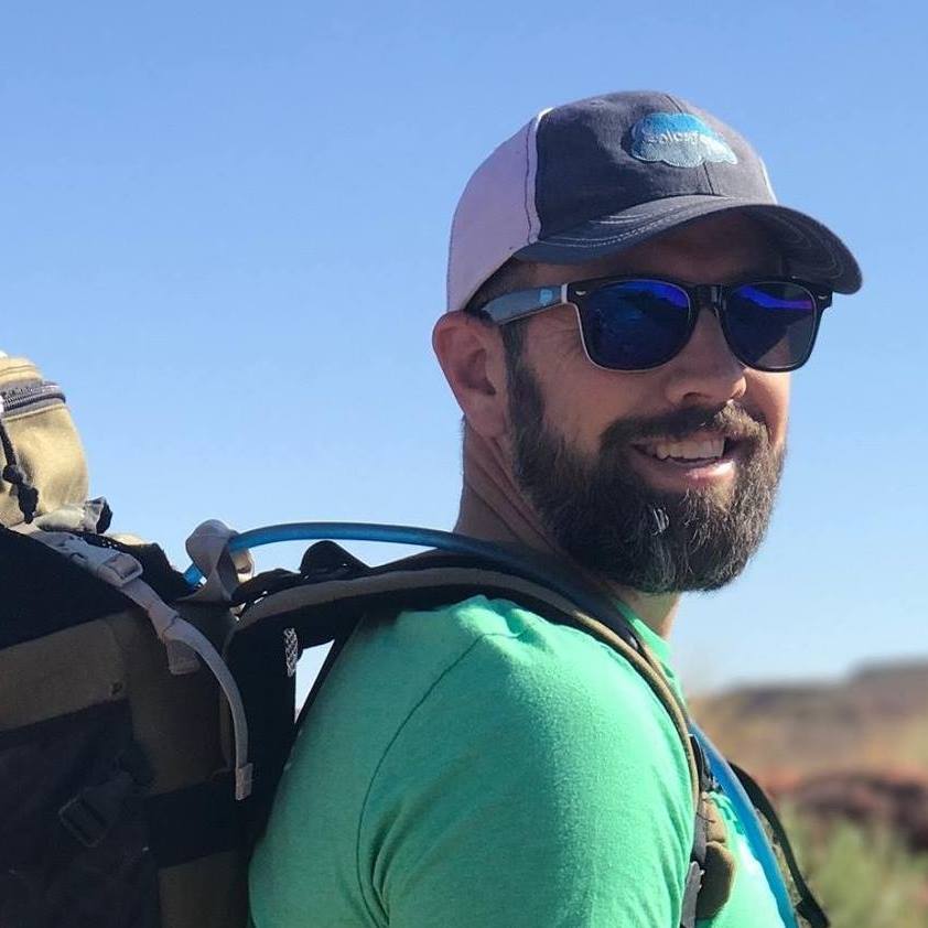 A headshot of artist Scott Barlow outdoors. He is weaing a hiking camelbak bag and sunglasses.