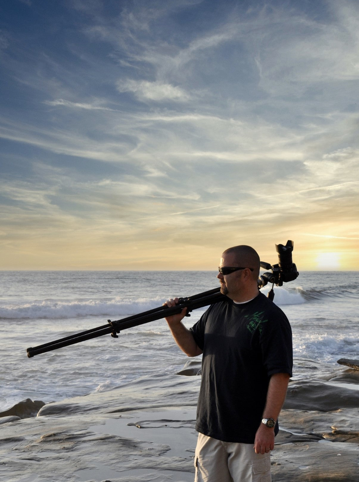 Artist Kelly Wade standing on a beach, holding his camera and tripod.