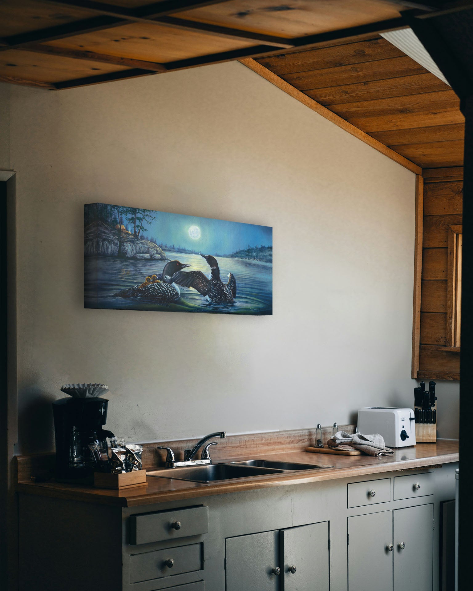 A painting hanging on a kitchen wall. A painting of two grown loons, with two baby loons on one of their backs. They are floating on a lake under the moonlight, with a campground on the land in the distance. Printed on canvas.