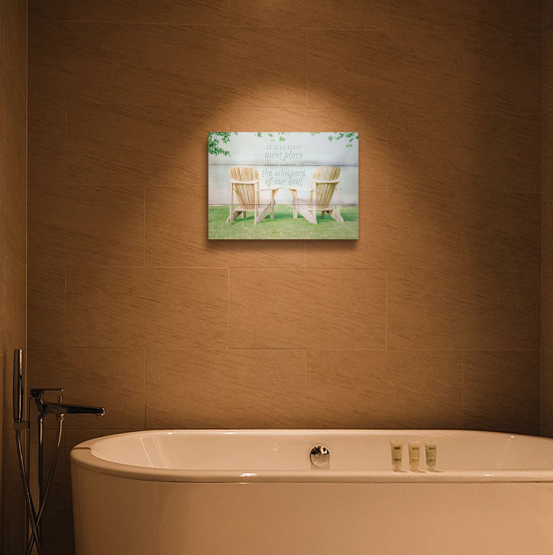 A photograph hanging on the wall of a bathroom. A photo of two wood lawn chairs, sitting on bright green grass, facing a lake. It is overlaid with the phrase, "It is in that quiet place that we hear the whispers of our soul." Printed on a box board.