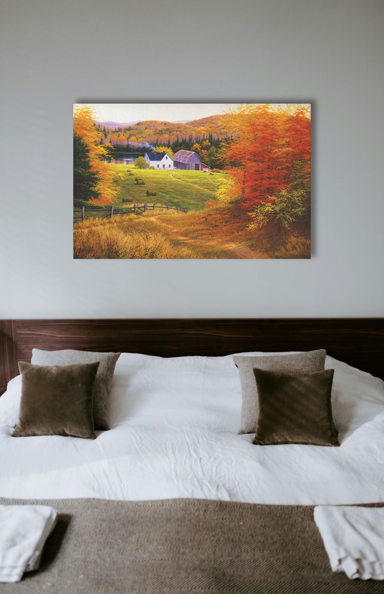 A painting hanging on a bedroom wall. A painting of a view looking down on a lakeside home and barn from a forest, featuring a few farm animals and hay bales. Printed on canvas.