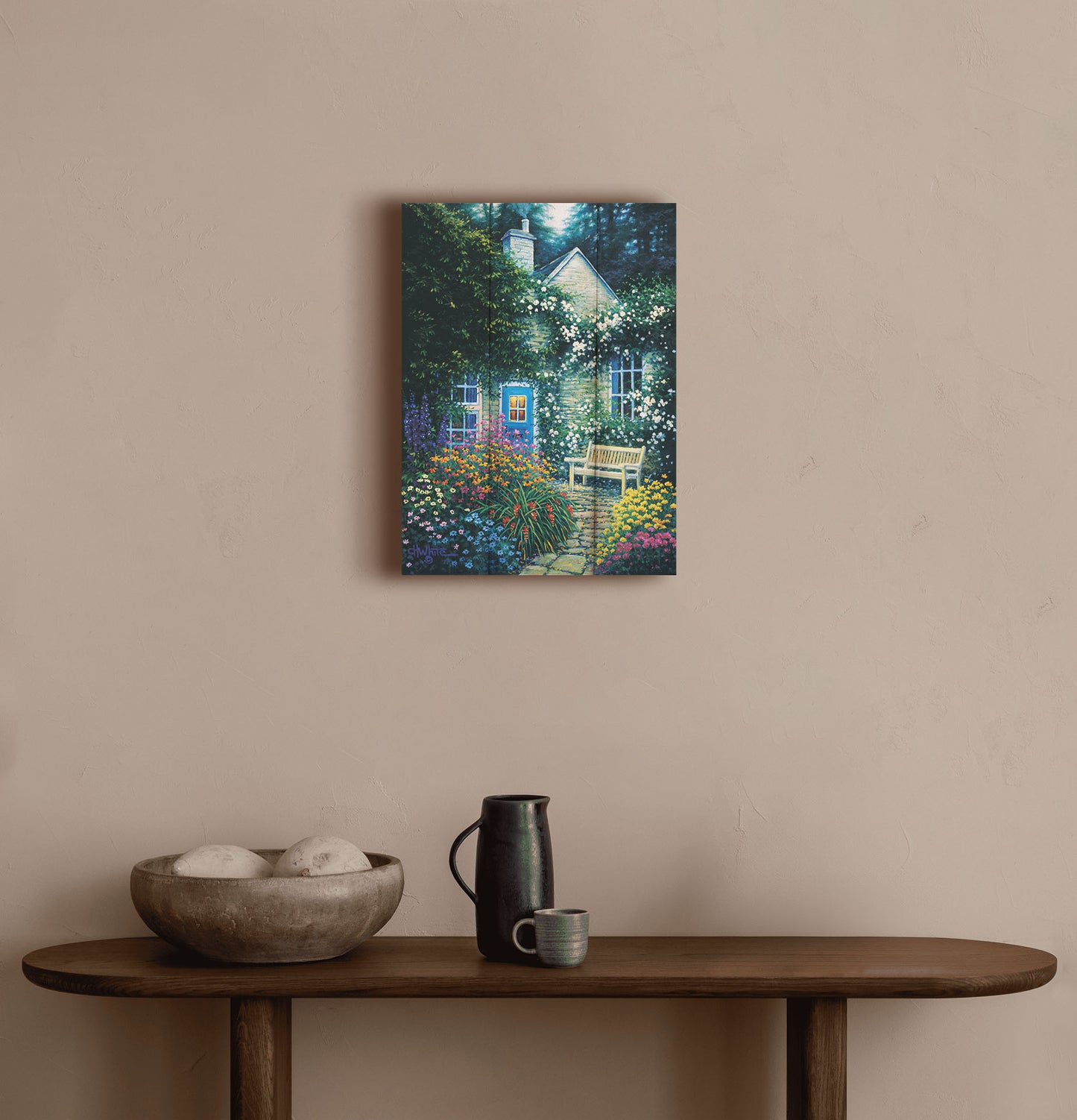 A painting hanging on a hallway wall. A painting of a cottage and stone path with a bench, all surrounded by colorful flower bushes and vines. Printed on a box board.