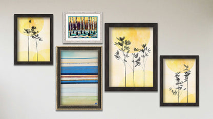 A collage of art in the designer Feeling Love package, including a series of three framed photos of plants silhouetted against yellow skies, a framed abstract geometric painting invoking boats and the shore, and a matted and framed painting of birch trees in winter against a yellow sky.