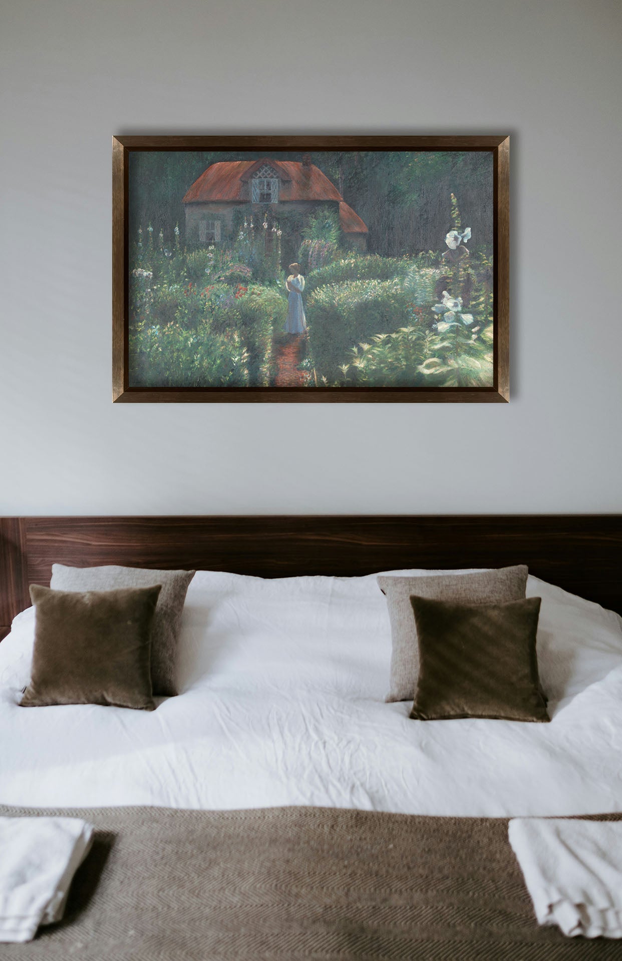 A typical bedroom with light walls and a cozy brown bed. A framed painting of a cottage surrounded by a garden hangs on the wall.